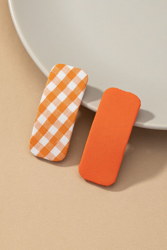 Two orange hair clips, one checkered and one solid, on a beige surface with a white plate in the background.