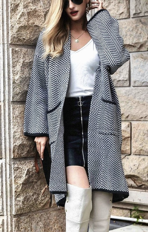 Person wearing a long black and white patterned coat over a white top and black skirt, standing against a stone wall.