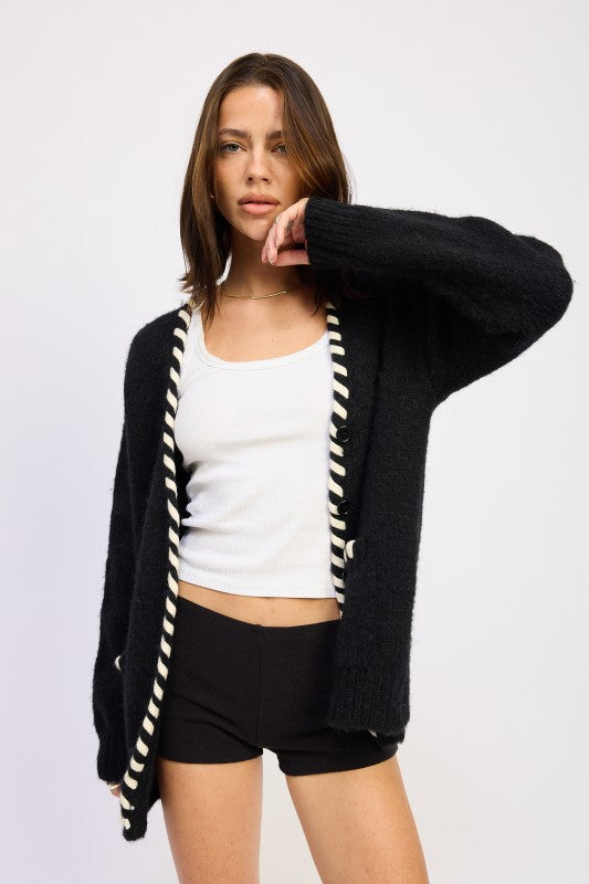 Woman wearing a black cardigan with white trim, white top, and black shorts on a light gray background