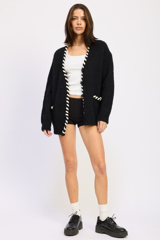 Woman wearing a black cardigan with white trim, white top, black shorts, and black shoes on a white background