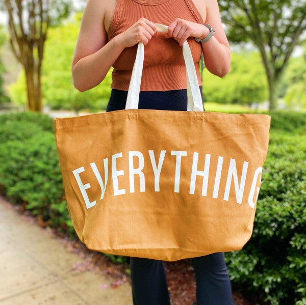 Person holding a mustard yellow tote bag with 'EVERYTHING' printed on it, outdoors.