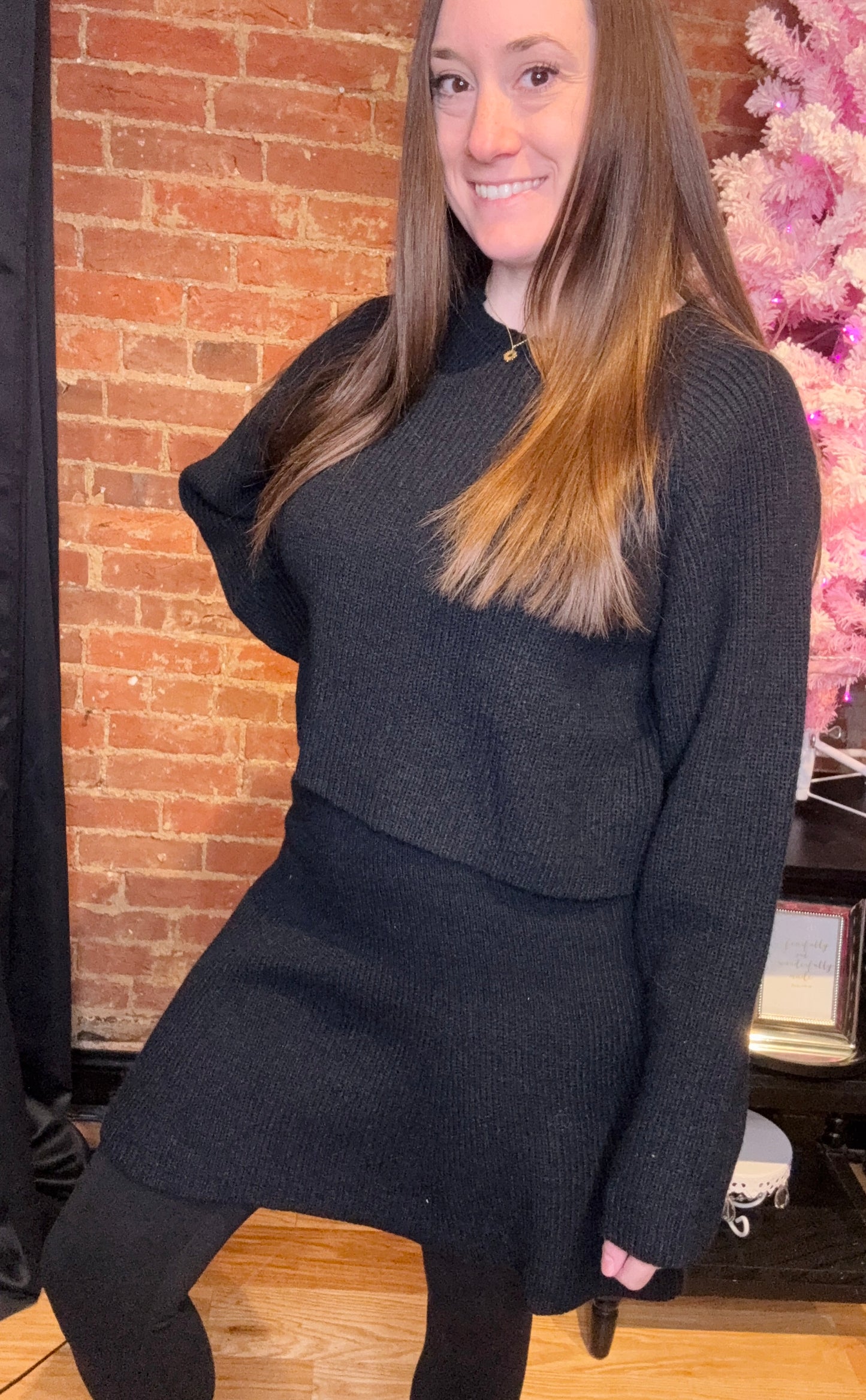 A woman standing in a bricked room wearing a black ribbed sweater and a mini skirt. She has long sleeves and is posing with one hand on her hip.