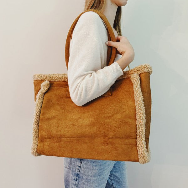 Person holding a brown tote bag with shearling trim against a plain background