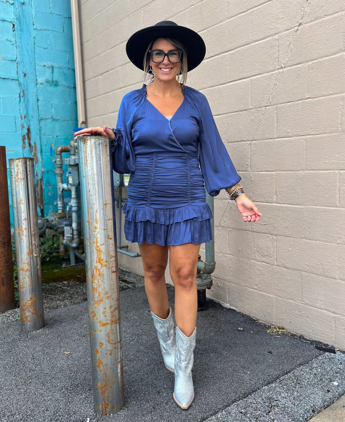 A person wearing a steel blue ruffled and shirred dress with long sleeves, paired with white boots and a wide-brimmed hat.