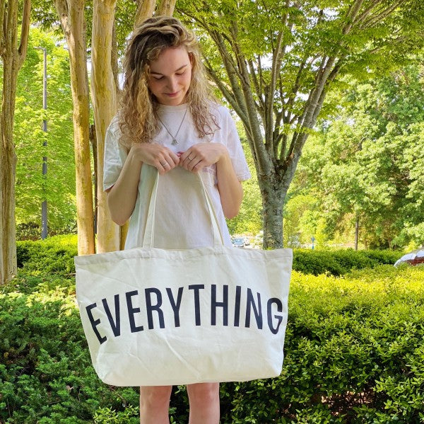 Person holding a tote bag with 'EVERYTHING' printed on it in an outdoor setting.