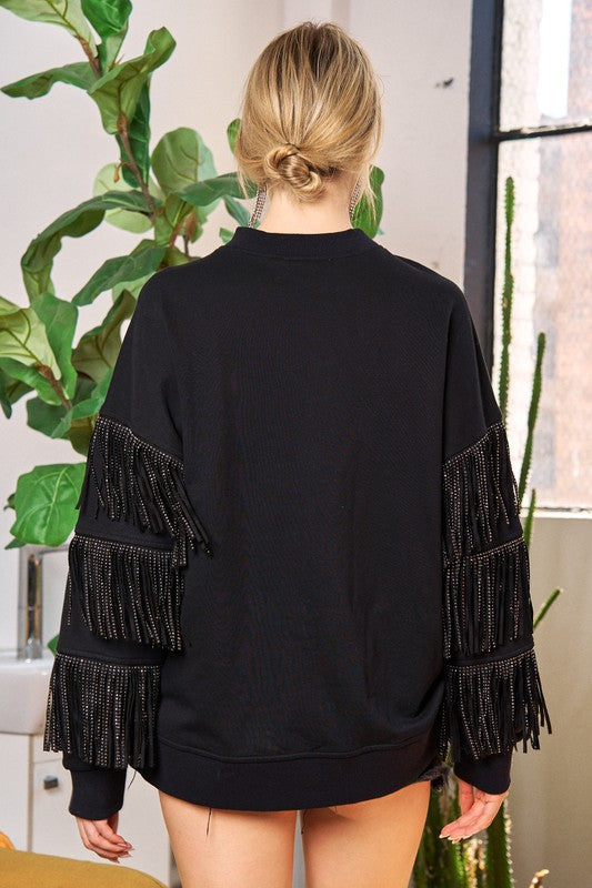 Person wearing a black sweater with fringe details in a room with plants.