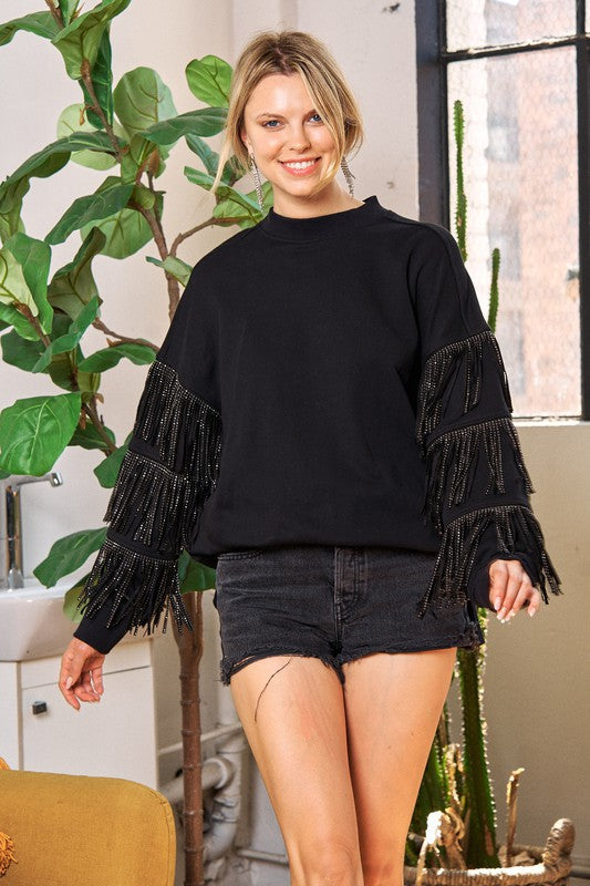 Woman wearing a black fringed sweater and denim shorts standing indoors with plants in the background.