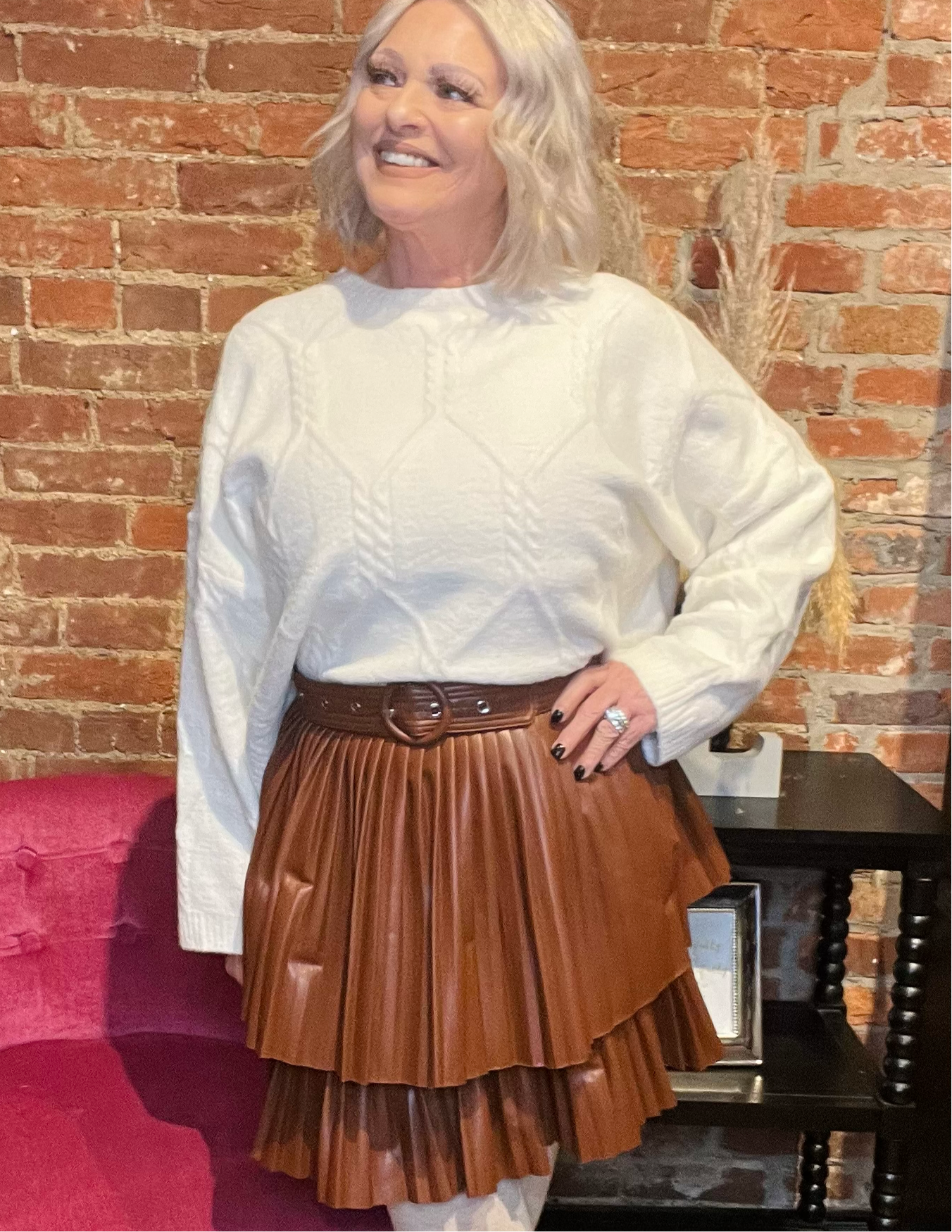 A woman standing in a studio setting, wearing a white belted pleated mini skirt and a white long-sleeve top with a crew neckline. The skirt has a brown color at the bottom, and the woman is posing with a smile.