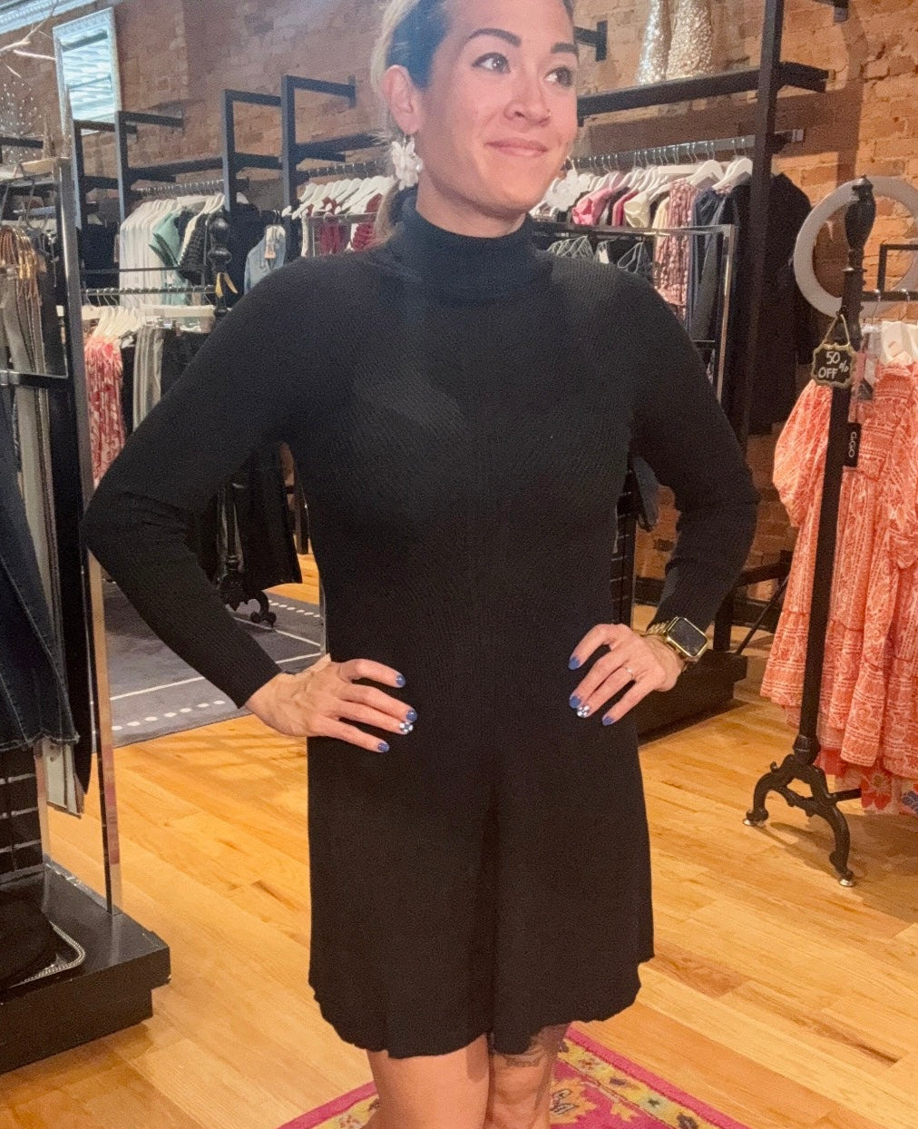 Woman wearing a black dress in a clothing store with racks of clothes in the background.