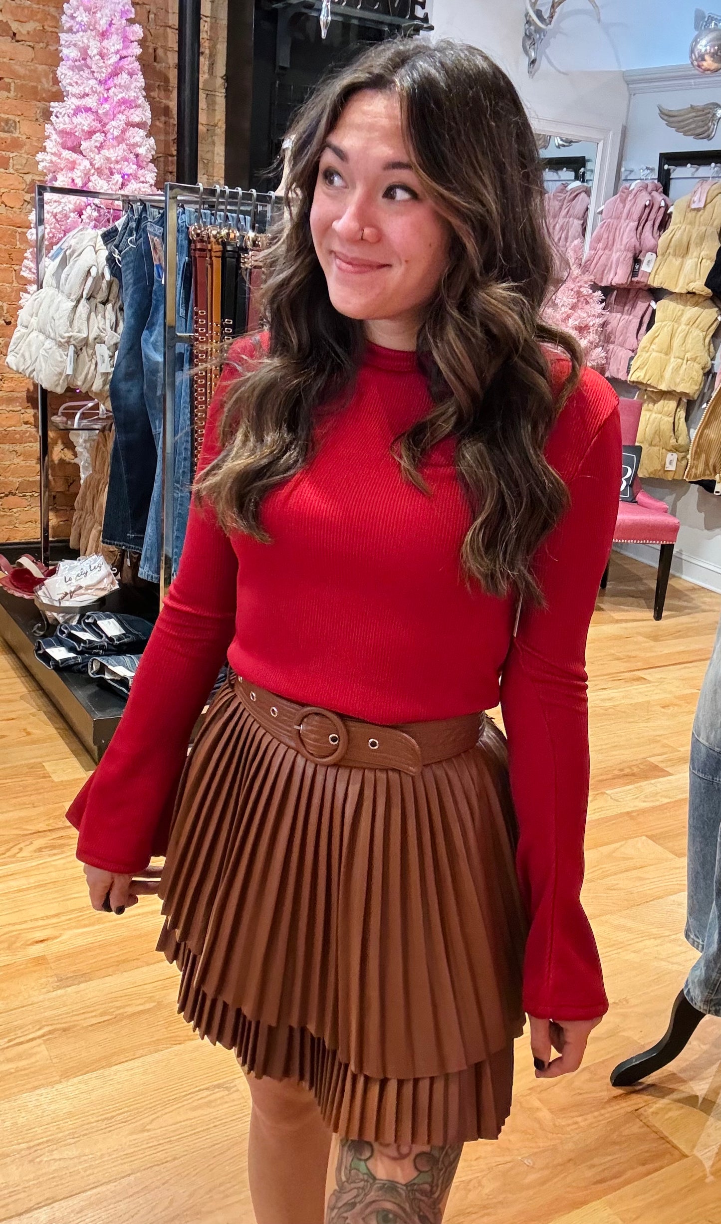 A woman standing in a studio setting, wearing a white belted pleated mini skirt and a white long-sleeve top with a crew neckline. The skirt has a brown color at the bottom, and the woman is posing with a smile.