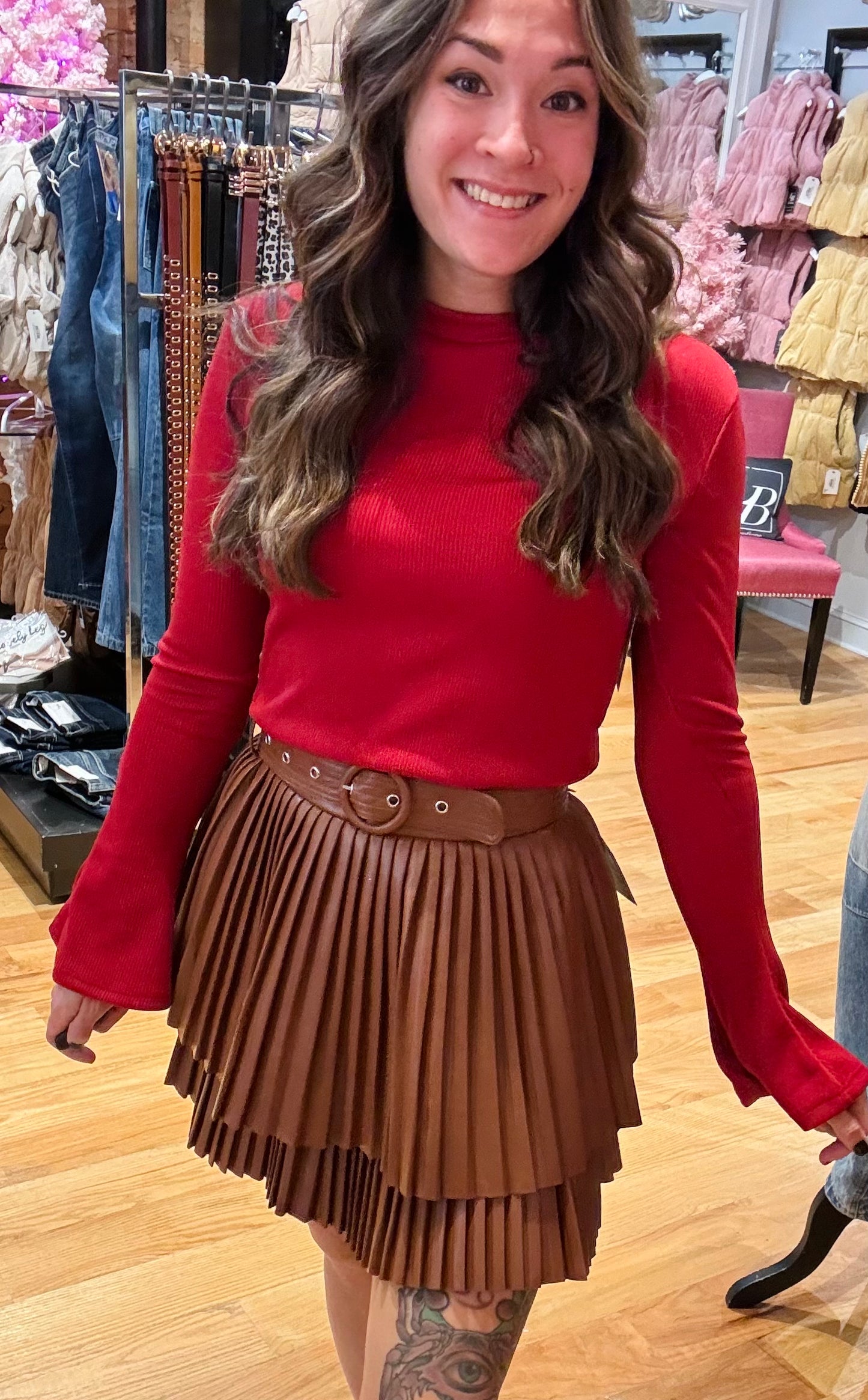 A woman standing in a studio setting, wearing a white belted pleated mini skirt and a white long-sleeve top with a crew neckline. The skirt has a brown color at the bottom, and the woman is posing with a smile.