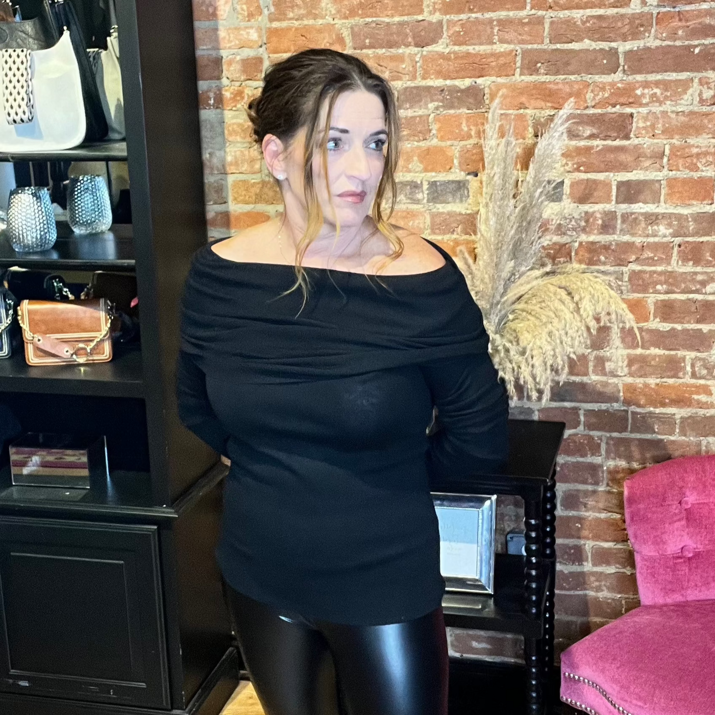 A woman standing in a salon chair wearing a black off-the-shoulder knit sweater top with long sleeves.