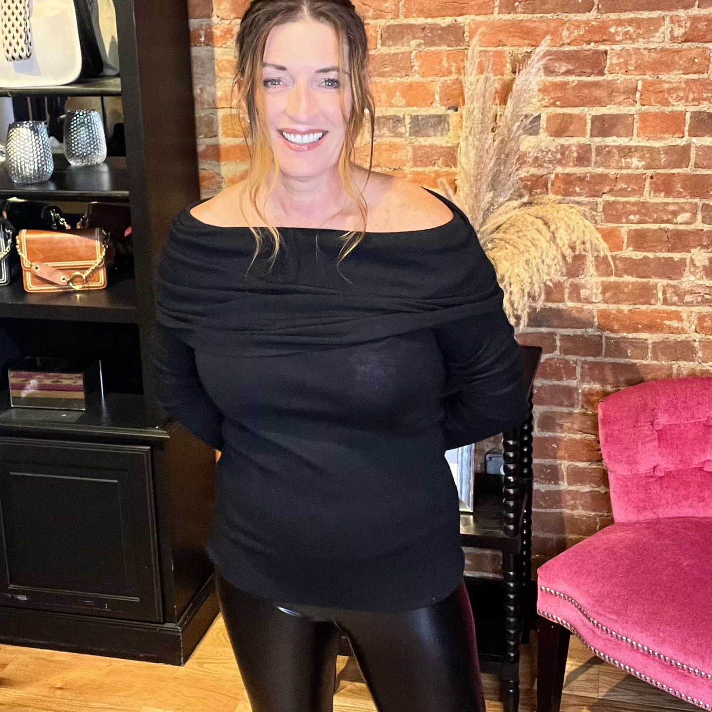 A woman standing in a salon chair wearing a black off-the-shoulder knit sweater top with long sleeves.