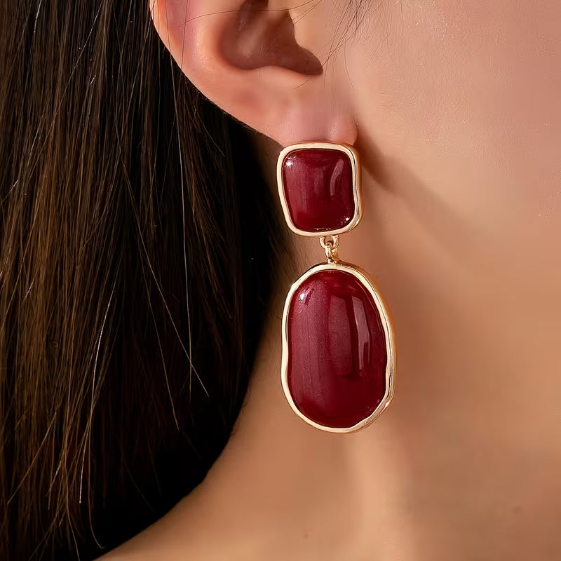 A close-up of a model's ear wearing a drop earring with a small square red stone and a larger irregular oval red stone, both set in gold-tone metal.