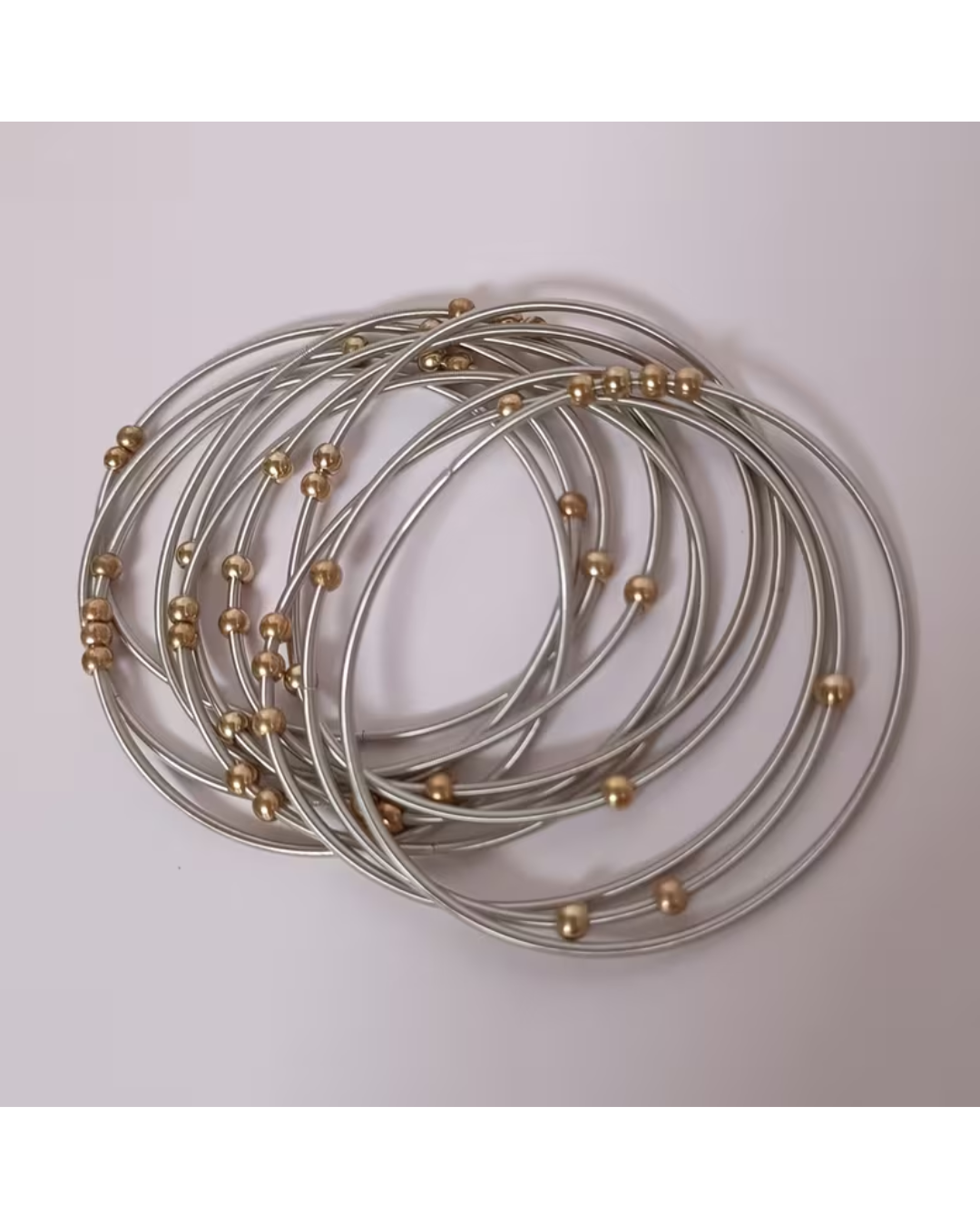 A set of thin silver-toned bangles with small metallic gold beads spaced along the bands, arranged in a loose coil on a plain light background.