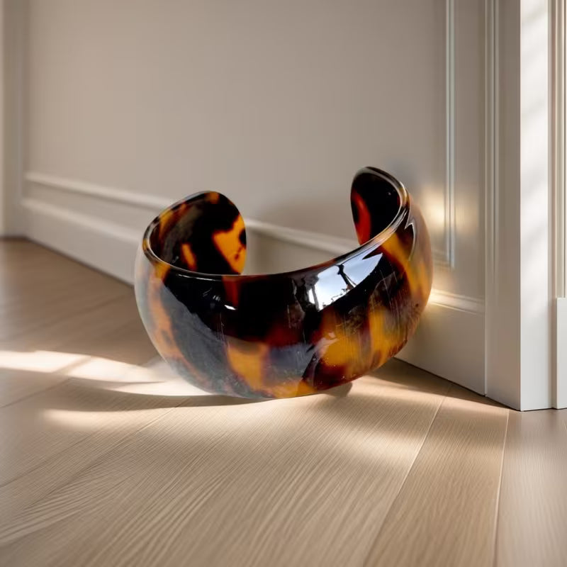 A glossy tortoiseshell cuff bracelet with a warm amber, orange, and black pattern, resting on a light wood floor next to a white door frame in soft sunlight.