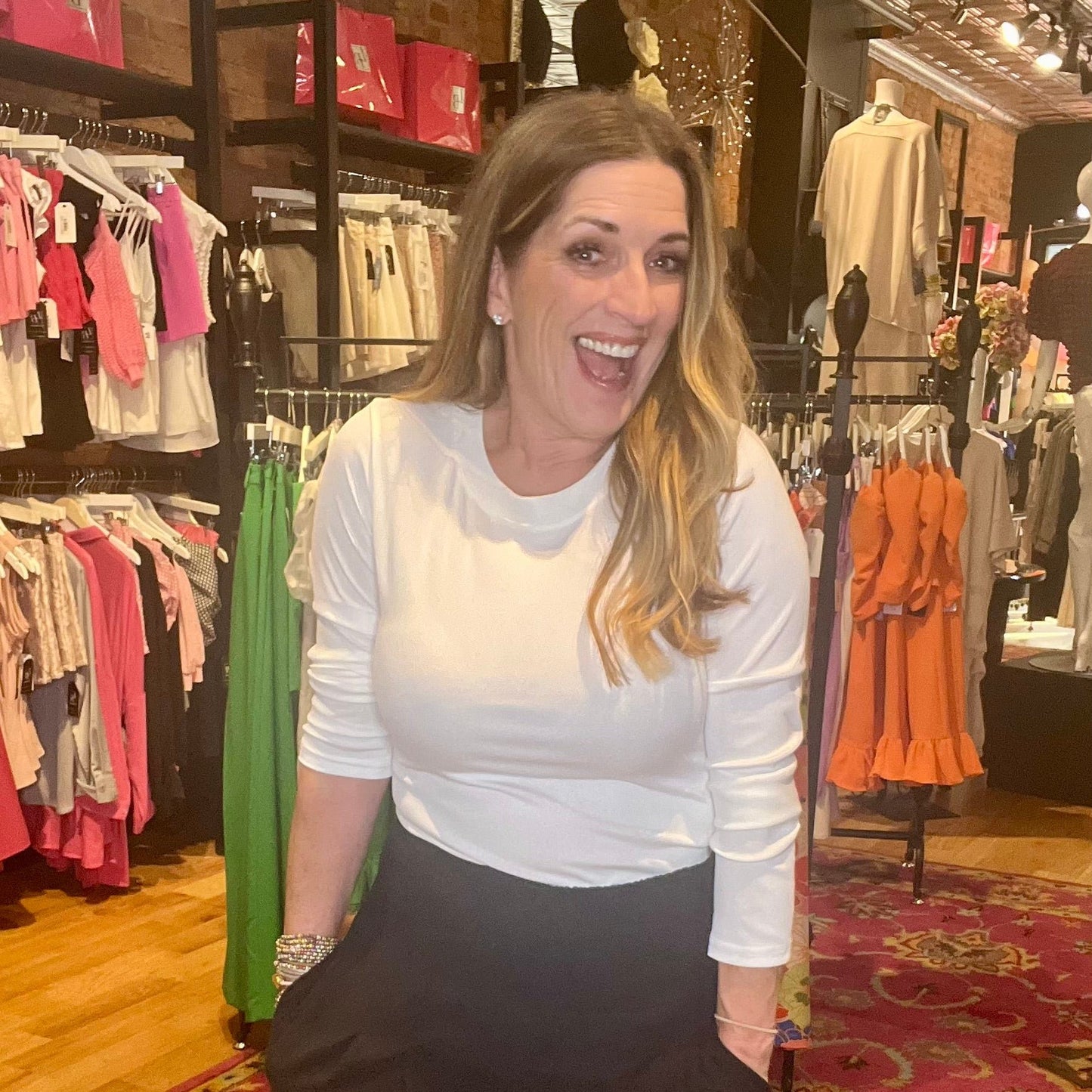 A woman standing in a retail environment, wearing a white ribbed long sleeve tee with a round neckline, and black skirt.