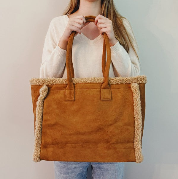 Person holding a brown tote bag with shearling trim against a plain background
