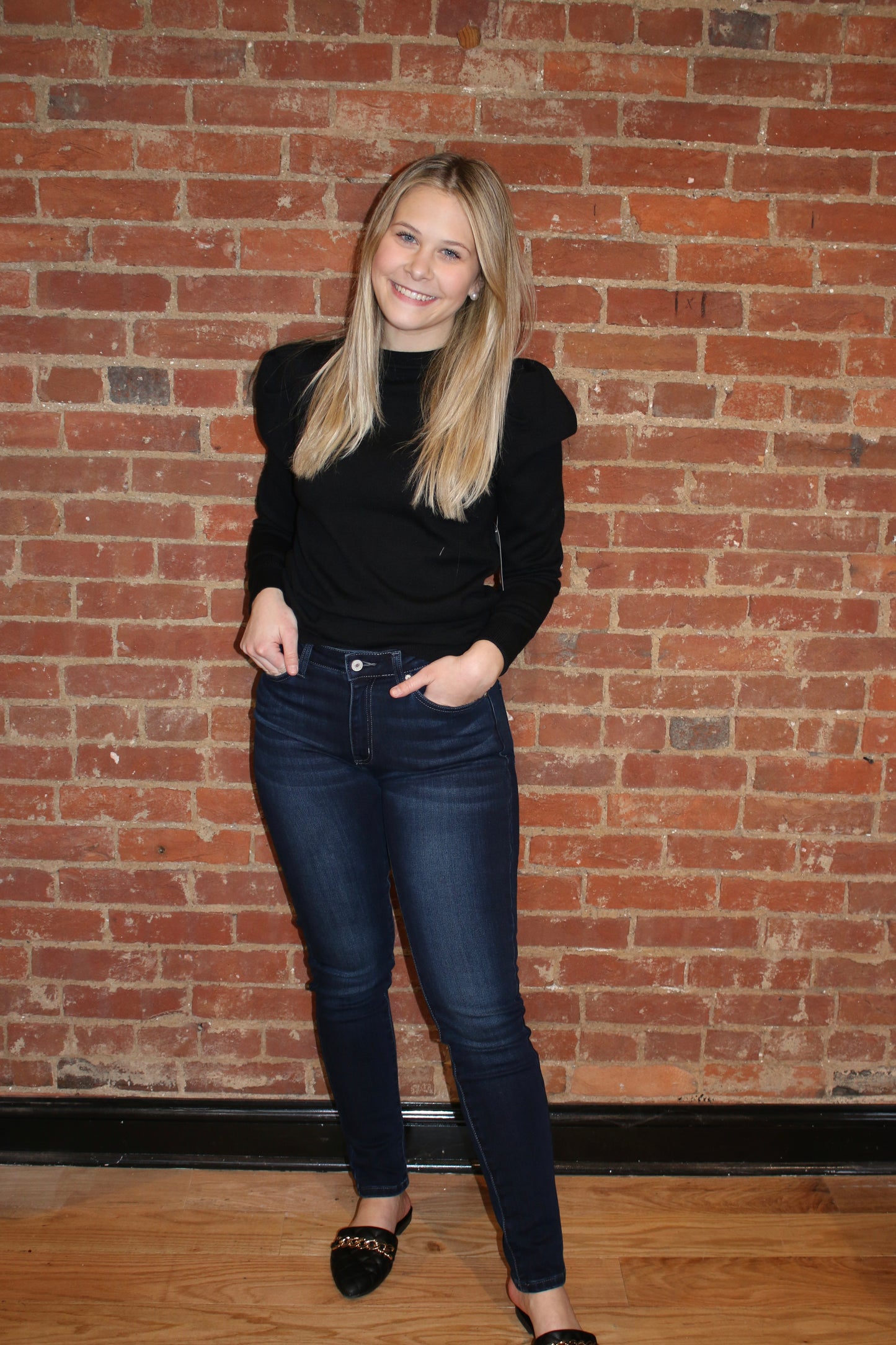 A person standing against a brick wall background, wearing dark wash skinny jeans and a black top.