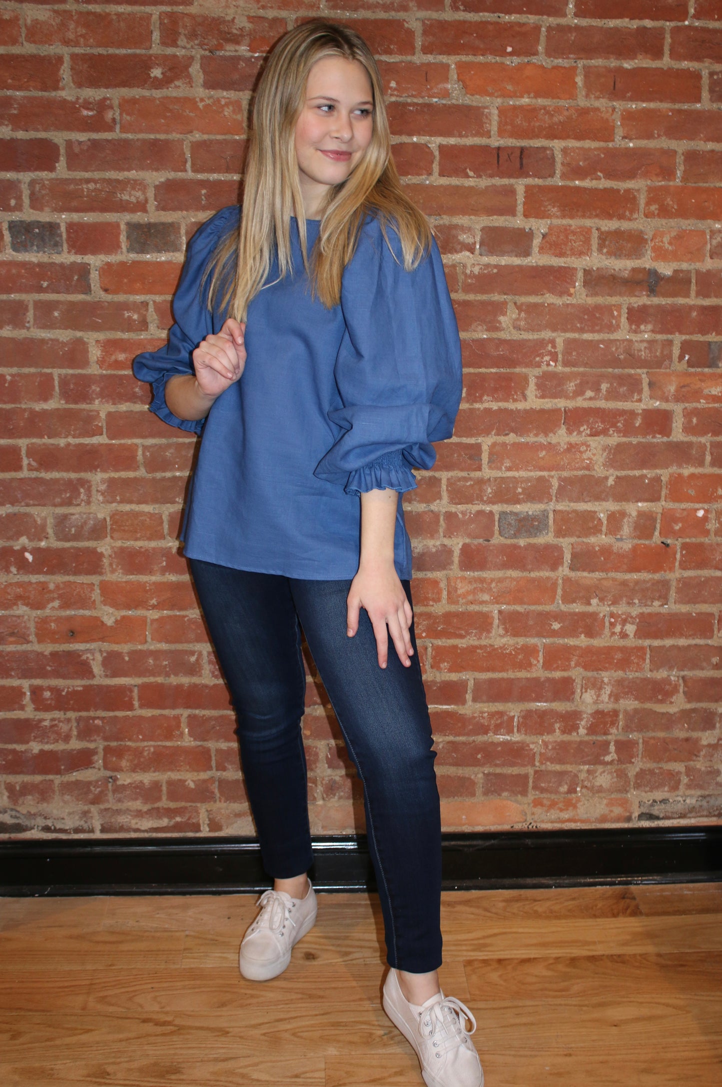 A person standing against a brick wall background, wearing dark wash skinny jeans and a black top.