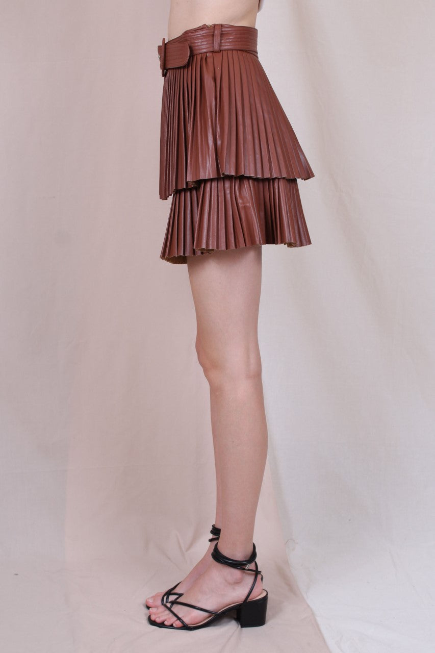 A woman standing in a studio setting, wearing a white belted pleated mini skirt and a white long-sleeve top with a crew neckline. The skirt has a brown color at the bottom, and the woman is posing with a smile.