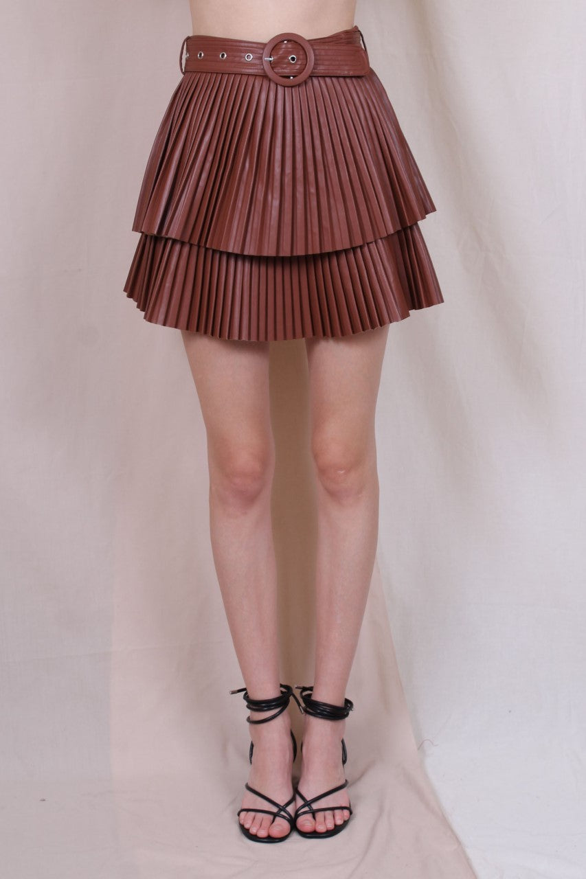 A woman standing in a studio setting, wearing a white belted pleated mini skirt and a white long-sleeve top with a crew neckline. The skirt has a brown color at the bottom, and the woman is posing with a smile.