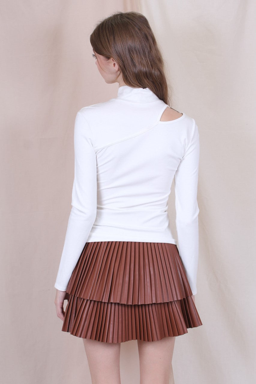 A woman standing in a studio setting, wearing a white belted pleated mini skirt and a white long-sleeve top with a crew neckline. The skirt has a brown color at the bottom, and the woman is posing with a smile.