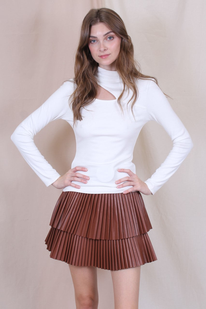 A woman standing in a studio setting, wearing a white belted pleated mini skirt and a white long-sleeve top with a crew neckline. The skirt has a brown color at the bottom, and the woman is posing with a smile.