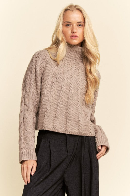 Woman wearing a beige cable knit sweater and black pants on a beige background