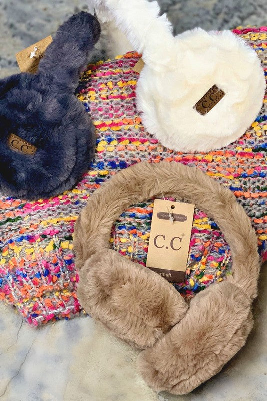 Pair of beige fur earmuffs with a visible brand tag on a colorful textured surface.