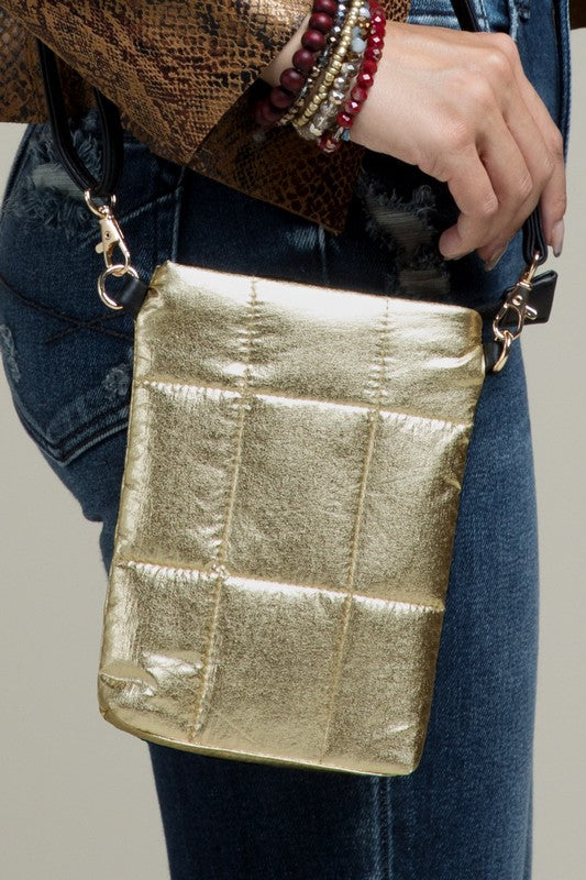 A metallic gold crossbody bag with a quilted design, featuring a faux leather strap and zipper closure, held by a person wearing blue jeans.