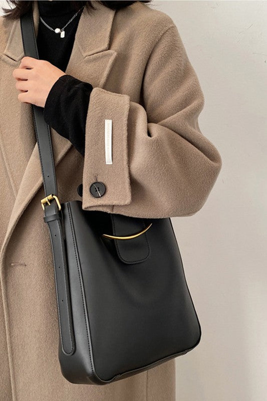 Person wearing a beige coat holding a black shoulder bag against a neutral background