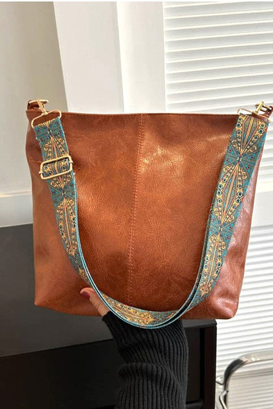 Brown leather handbag with decorative strap held by a person against a neutral background