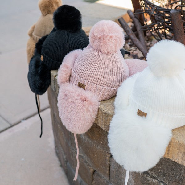 Three knit hats with pom-poms in black, pink, and white on a stone ledge.