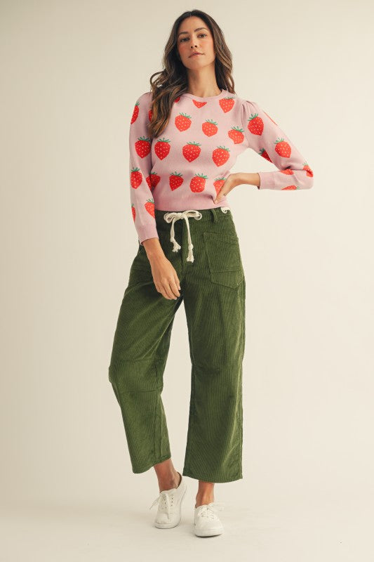 Woman wearing a pink sweater with red strawberry pattern and green pants on a beige background