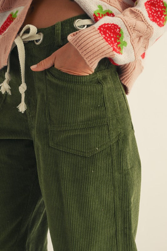 Person wearing green corduroy pants with a pink sweater with strawberry pattern on sleeves.