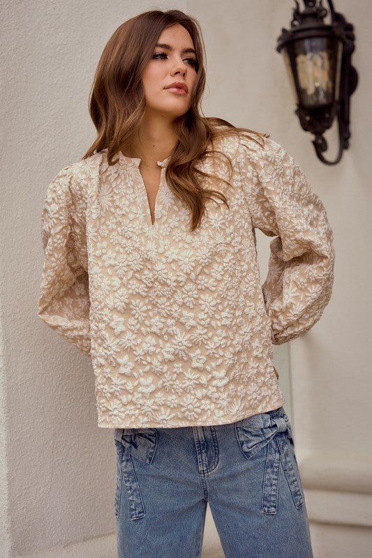 Woman wearing a beige floral blouse and blue jeans against a neutral wall.