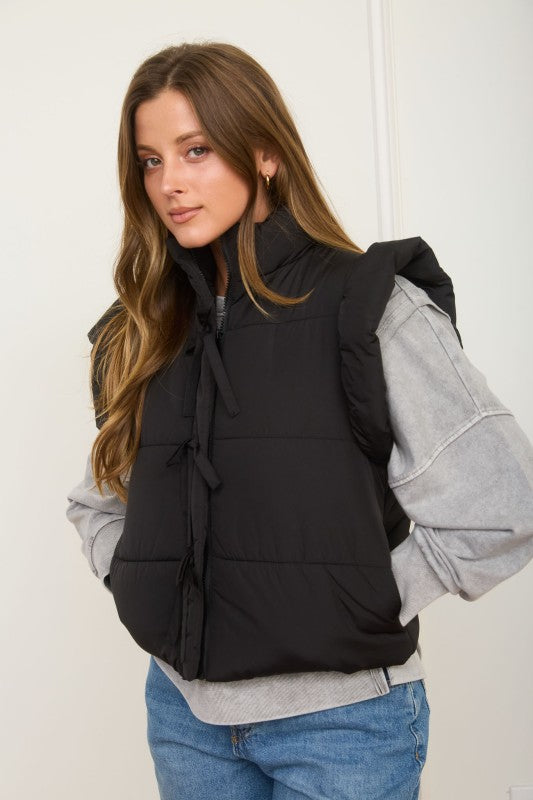 Woman wearing a black puffer vest over a gray sweater with a white background