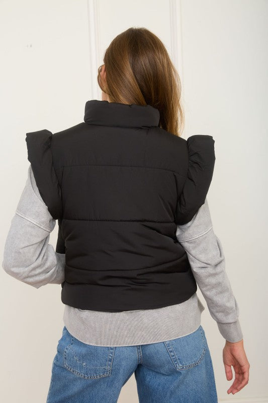 Person wearing a black puffer vest over a gray sweater and blue jeans on a white background
