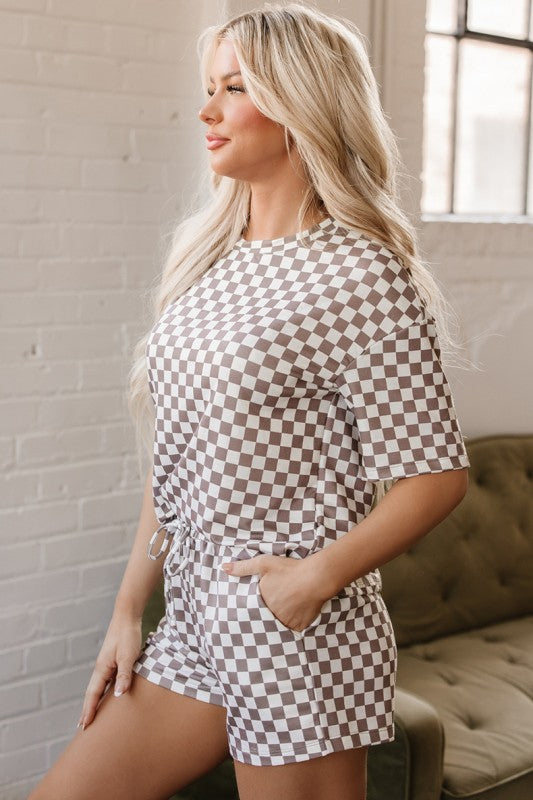 Woman wearing a checkered outfit standing in a room with a brick wall and window.