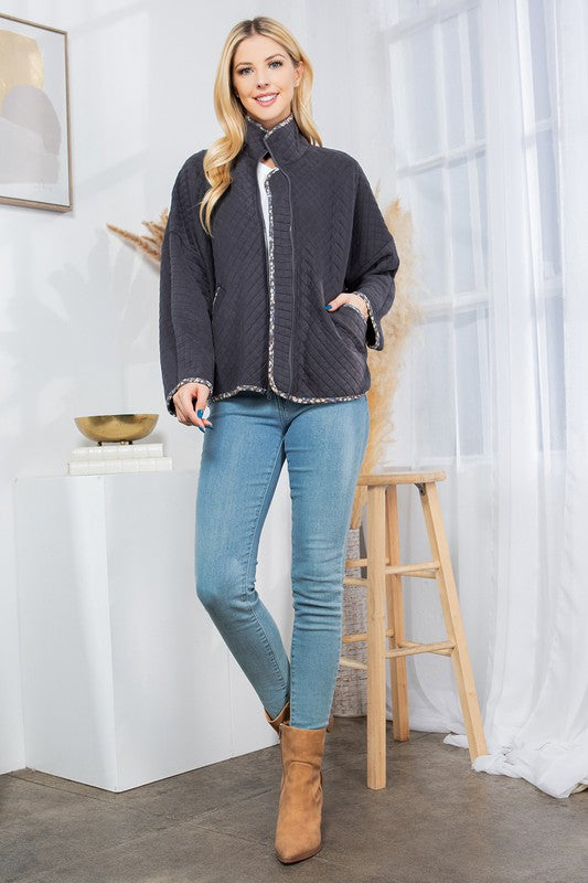 Woman wearing a dark gray jacket, light blue jeans, and brown boots in a room with white walls and a wooden stool.