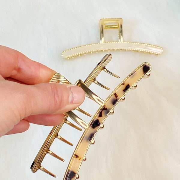Gold hair clip with tortoiseshell pattern held by a hand on a white background