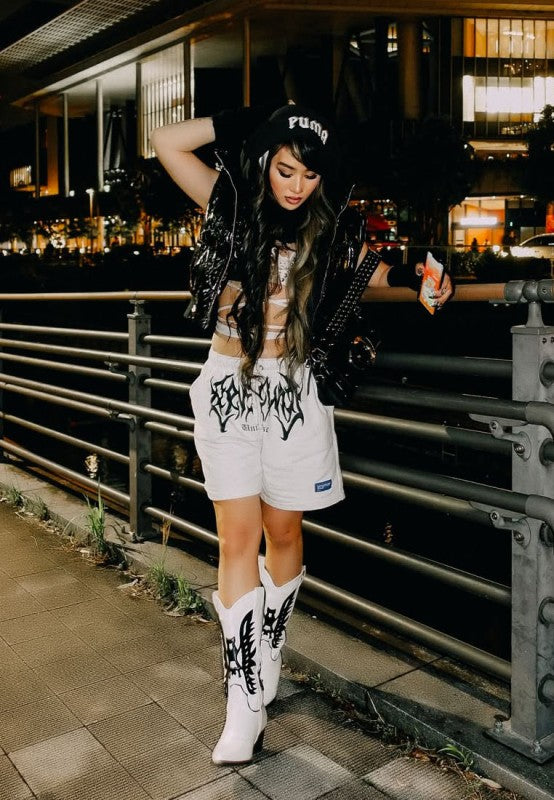 Person wearing a black jacket, white shorts with graphic design, and white boots in an urban setting at night.
