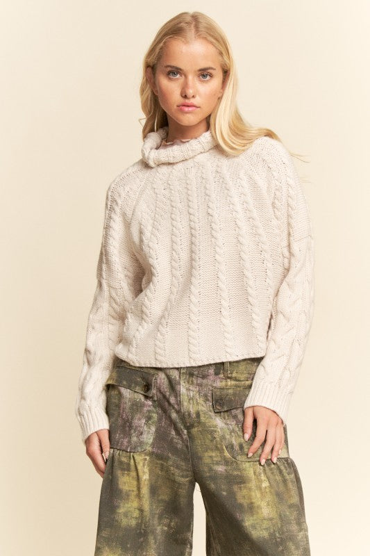 Woman wearing a cream cable knit sweater and patterned pants against a beige background