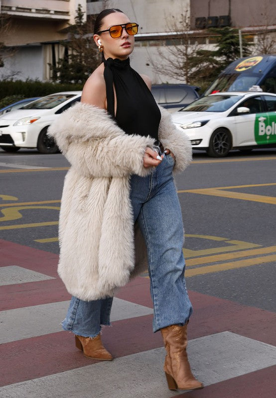 Person wearing a fluffy beige coat, black top, blue jeans, and brown boots on a city street.