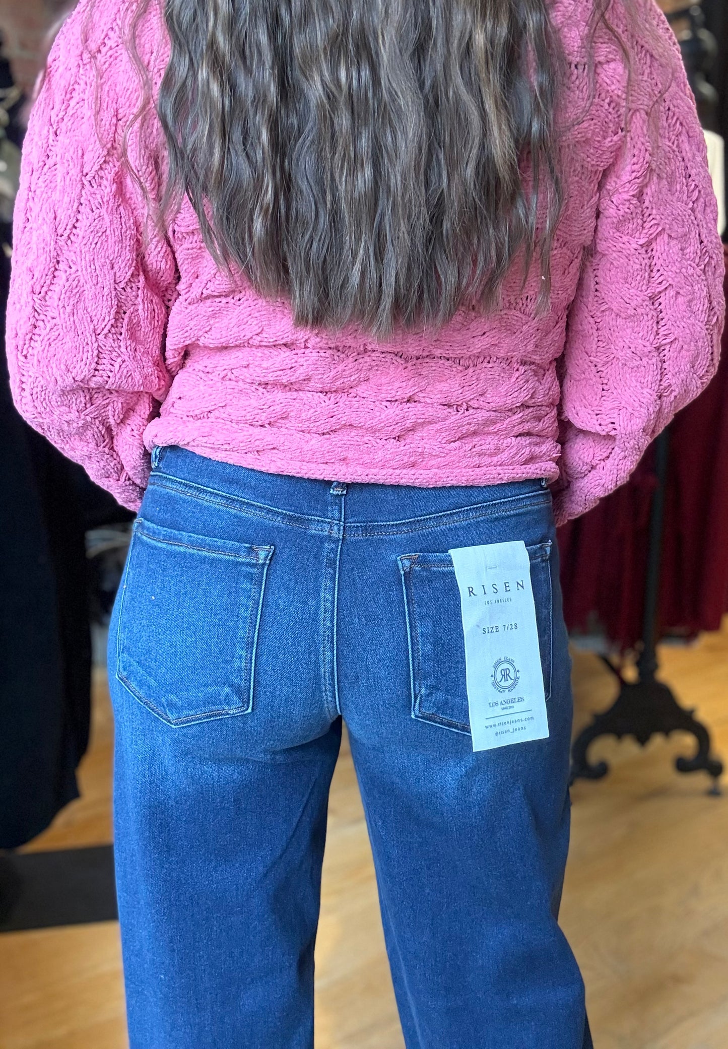 Person wearing a pink sweater and blue jeans with a visible tag, standing indoors.