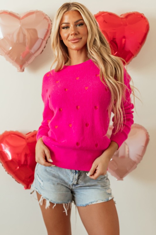Woman wearing a pink sweater with heart patterns and denim shorts, surrounded by heart-shaped balloons.
