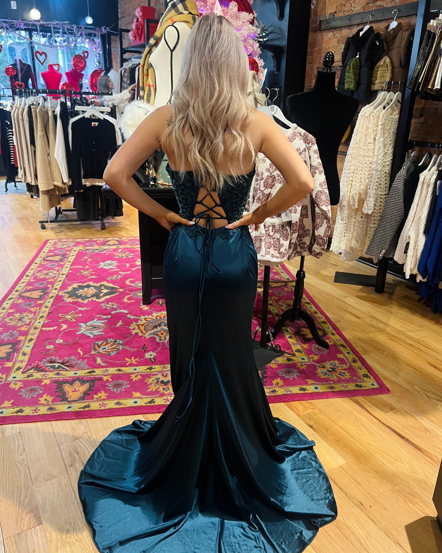 Woman in a teal evening gown standing in a clothing store with racks of clothes and a colorful rug on the floor.