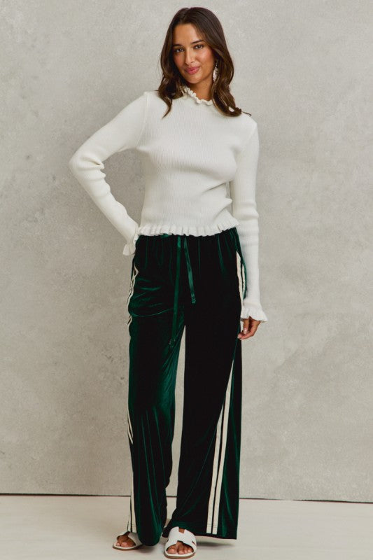 Woman wearing a white sweater and green pants with white stripes against a plain background