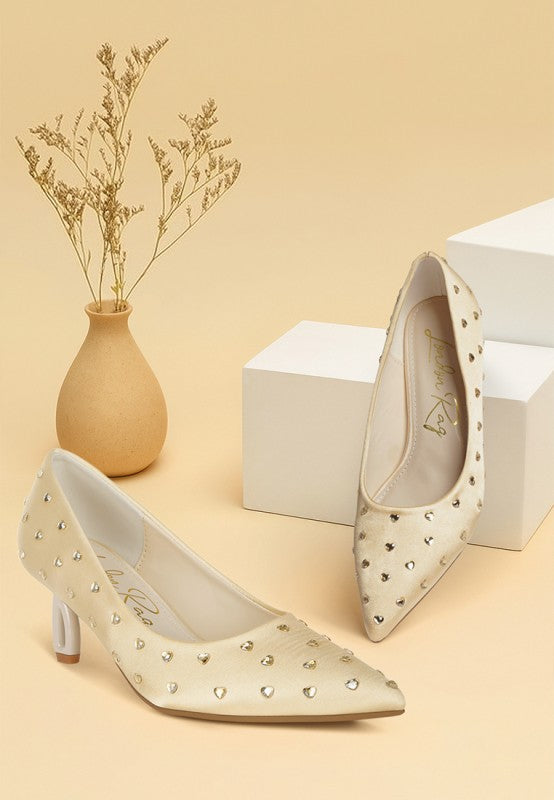 Pair of beige high-heeled shoes with heart details on a beige background with a vase and box.